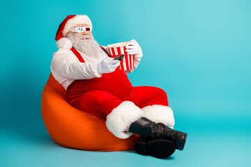Funny Santa relaxes on orange beanbag with popcorn holding a remote against a blue background in a festive holiday scene