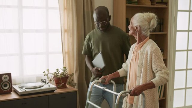 Tilt up shot of white haired senior woman using walker slowly taking steps at home and smiling with caring Black man as social worker assisting, copy space