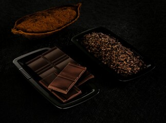 Cacao pods and finely chopped cocoa beans are elegantly displayed alongside smooth artisanal chocolate bars on a sleek black surface. The scene captures the essence of chocolate creation