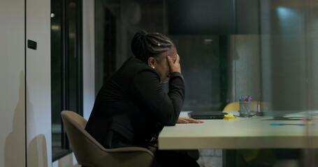 African American businesswoman sits in silence at office table with hand over face, processing...