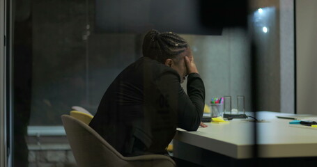 African American businesswoman sits in silence at office table with hand over face, processing emotional stress and fatigue after long and intense workday