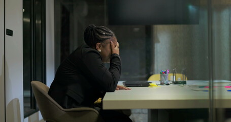 African American businesswoman sits in silence at office table with hand over face, processing...