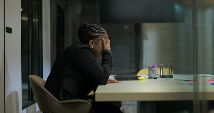 African American businesswoman sits in silence at office table with hand over face, processing emotional stress and fatigue after long and intense workday