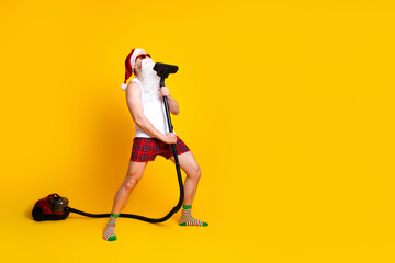 Santa in a white tank top plaid shorts and striped socks cleans with a vacuum against a bright...