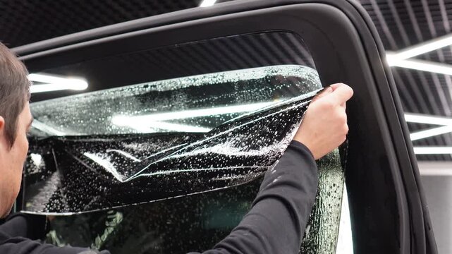 Applying tinting foil on a car window in the tuning studio. Male car service worker glue window tinting in luxury car detailing car repair the concept of love for cars and protection. Wrapping auto. 
