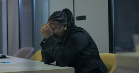 African American businesswoman sits with face buried in hands at office desk, struggling with...