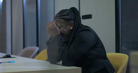 African American businesswoman sits with face buried in hands at office desk, struggling with...