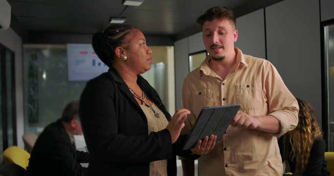 African American businesswoman explaining tablet data to young male colleague in office, active teamwork near conference table with charts on screen