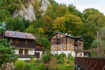 Old wooden mountain houses stand at the base of towering limestone cliffs surrounded by colorful foliage. The blend of rustic architecture and natural rock scenery evokes rural lifestyle and mountain 