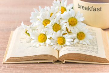 Summer still life; bouquet of white daisy or chamomile flowers on a book and cup of coffee on a wooden table; close up