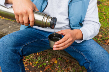 Fototapeta premium A close-up view of hands pouring a hot beverage from a stainless steel thermos into a small cup. Casual outdoor clothing and fallen leaves emphasize travel, hiking, warmth, and autumn comfort in natur
