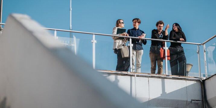 Four colleagues stand on a glass-railed rooftop terrace, chatting and sharing ideas under a clear blue sky. A candid moment of teamwork and casual business interaction.