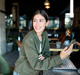 Woman smiling, holding coffee while working on a laptop and using a mobile phone  in a cafe