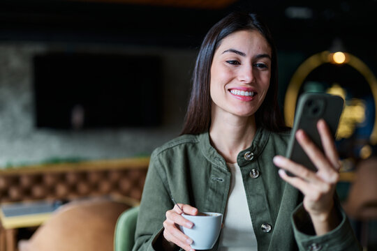 Portrait of a young woman girl  sitting in cafeteria or coffee shop drinking coffee or cappuccino and using a smartphone mobile phone  in the city and having fun