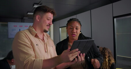 African American businesswoman smiling at digital tablet with young male colleague, collaborative moment in modern office during team meeting