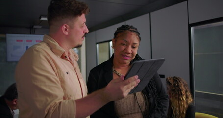 African American businesswoman smiling at digital tablet with young male colleague, collaborative moment in modern office during team meeting