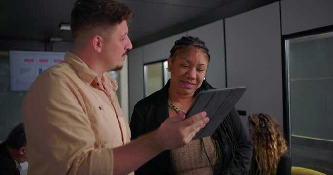 African American businesswoman smiling at digital tablet with young male colleague, collaborative moment in modern office during team meeting