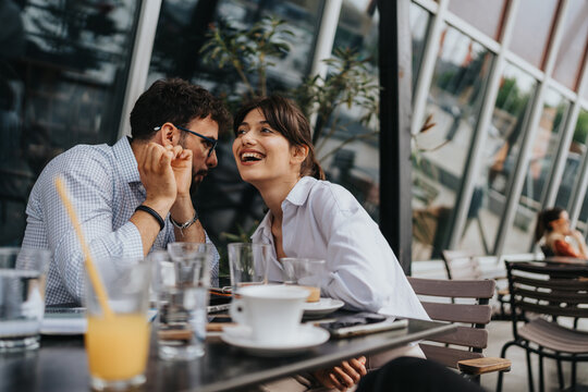 A cheerful couple shares laughter and conversation while seated at a cafe outdoors, surrounded by a lively atmosphere and modern architecture.