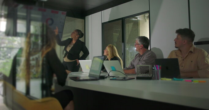 African American businesswoman presenting in office meeting while diverse team listens attentively in modern conference room with digital chart display