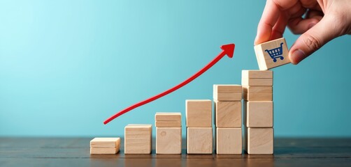 Hand places wooden block with shopping cart icon on top of stack. Wooden cubes form a rising graph. Red arrow indicates upward trend. Business growth concept on wooden table against blue background.