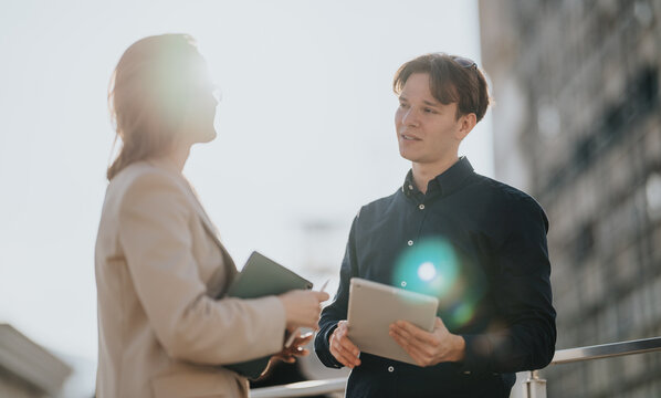 Two young business associates converse outside, one holding a tablet while the other carries a notebook. - Powered by Adobe