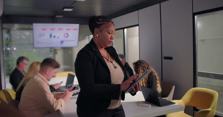 African American businesswoman standing in office using digital tablet, focused on work while colleagues collaborate at conference table in background