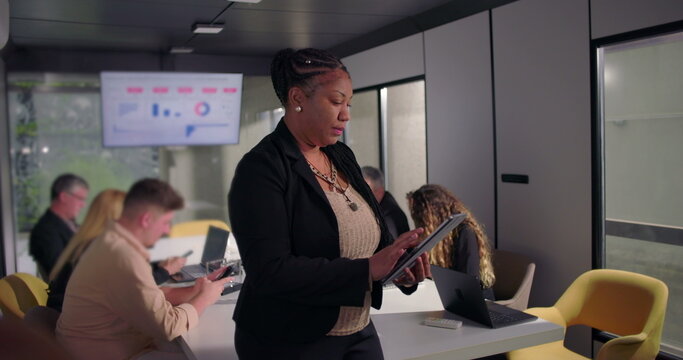 African American businesswoman standing in office using digital tablet, focused on work while colleagues collaborate at conference table in background