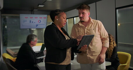 African American businesswoman showing tablet to young male colleague during office meeting, teamwork and analysis in front of large data display