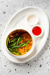 A warm bowl of beef pho or Asian noodle soup with snap peas, green onions, and fresh herbs, served with sriracha and cream in side dishes