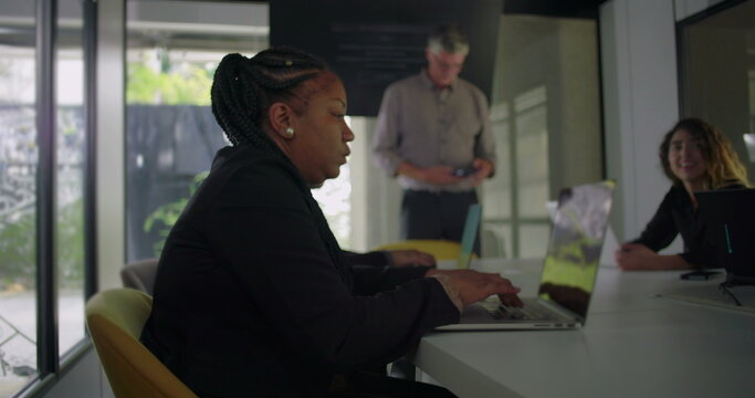 African American businesswoman typing during office meeting while colleagues collaborate in modern conference room with laptops and large windows - Powered by Adobe