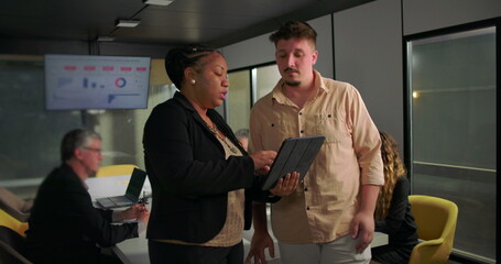 African American businesswoman showing tablet to young male colleague during office meeting, teamwork and analysis in front of large data display