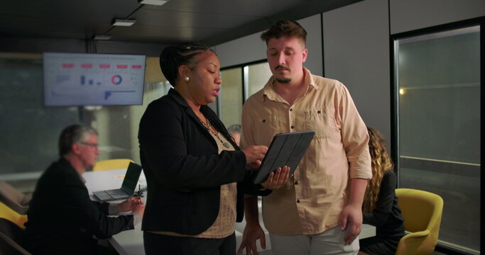 African American businesswoman showing tablet to young male colleague during office meeting, teamwork and analysis in front of large data display