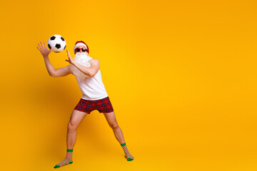 Santa Claus plays soccer in a funny home setting wearing a white tank top red plaid shorts and striped socks on a bright yellow background