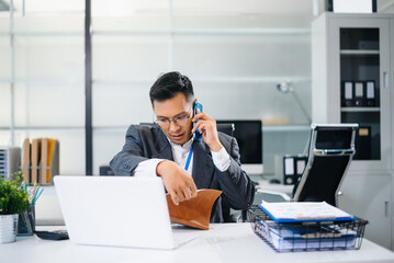 Asian businessman talking on smartphone while working on laptop at modern office desk, symbolizing business communication and corporate success.