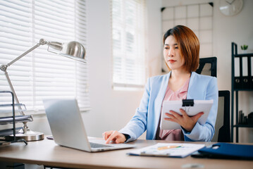 Asian businesswoman reviewing report with clipboard and laptop in bright office. Perfect for business, consulting, analysis, productivity, and corporate planning themes.