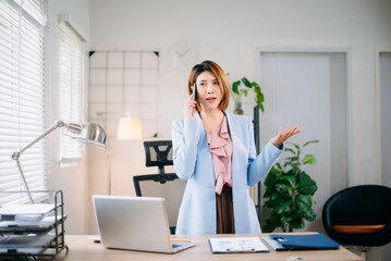 Confident Asian businesswoman talking on phone in bright modern office, working with laptop and financial reports. Perfect for business, consulting, leadership, and teamwork themes.