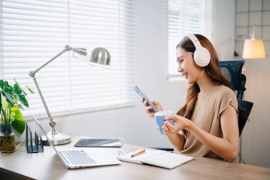 Asian woman working from home with laptop and headphones, smiling while using smartphone. and modern lifestyle themes.