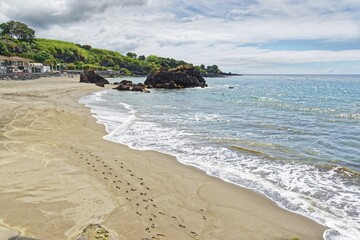 A quiet beach with footprints in the sand, gentle waves, clear water and rocks in the background under a partly cloudy sky, small town of Vila Franca do Campo, Sao Miguel, Azores, Portugal