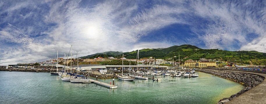 A picturesque harbour with yachts and boats, surrounded by buildings, mountains and a cloudy sky with sun, all in an impressive panoramic shot, small town of Vila Franca do Campo, Sao Miguel, Azores, Portugal