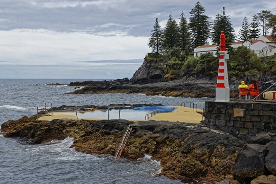 Small lighthouse and seawater swimming pool on the rocky coast under a cloudy sky, Caloura fishing village, Ribeira Ch&atilde;, Sao Miguel Island, Azores, Portugal
