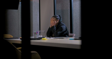 African American businesswoman sits at conference table with face in hands, seeking relief from...