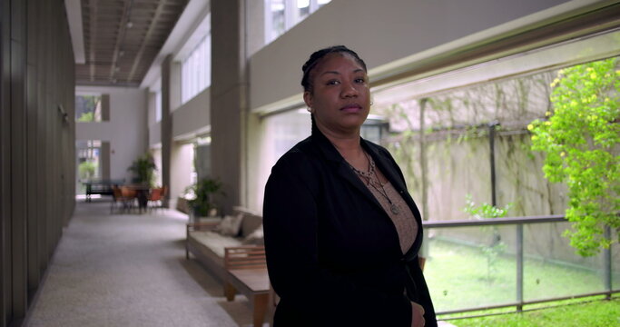 African American professional woman standing confidently in modern office hallway, looking at camera with calm expression beside large glass windows and greenery outside