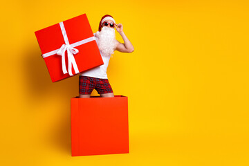 Santa Claus with a large red gift box wears sunglasses and plaid shorts in a bright yellow studio ready for festive gifts and holiday celebrations