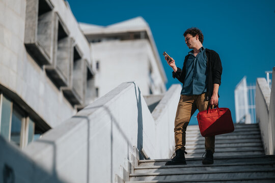 A young professional stands on concrete stairs outdoors, checking a smartphone while carrying a red bag. - Powered by Adobe