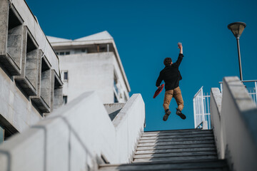 A young man in casual clothes leaps mid-air on concrete stairs outdoors. Bright blue sky, modern...
