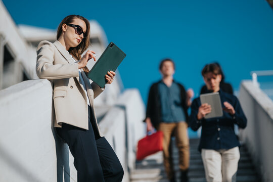 A stylish professional woman on a sunny day uses a tablet, with two teammates in the background, symbolizing teamwork, remote work, and modern business on the move.