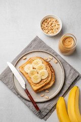 Peanut butter toast and banana in a plate on a light background with nuts, jar of spread and knife.