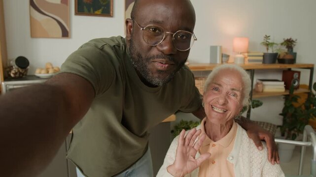 POV shot of Black man as social worker taking selfie with white haired woman at home both smiling while talking to camera, woman waving hello