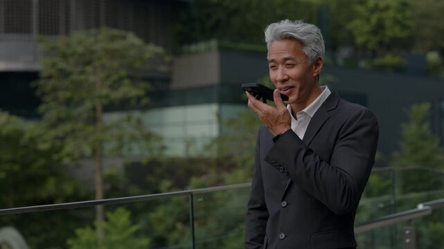 Portrait of grey haired Asian entrepreneur records audio message on mobile phone on office glass terrace. Japanese manager talks in voice chat via smartphone in downtown block