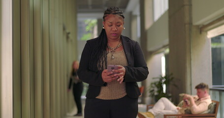 African American woman using smartphone while walking down office hallway, focused on screen with seated coworkers visible in background
